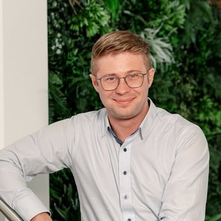 Professionelles Porträt eines Mannes in einem Hemd Der Mann trägt ein hellblaues Hemd und hat eine entspannte Haltung, während er vor einer lebendigen Pflanzenkulisse posiert.