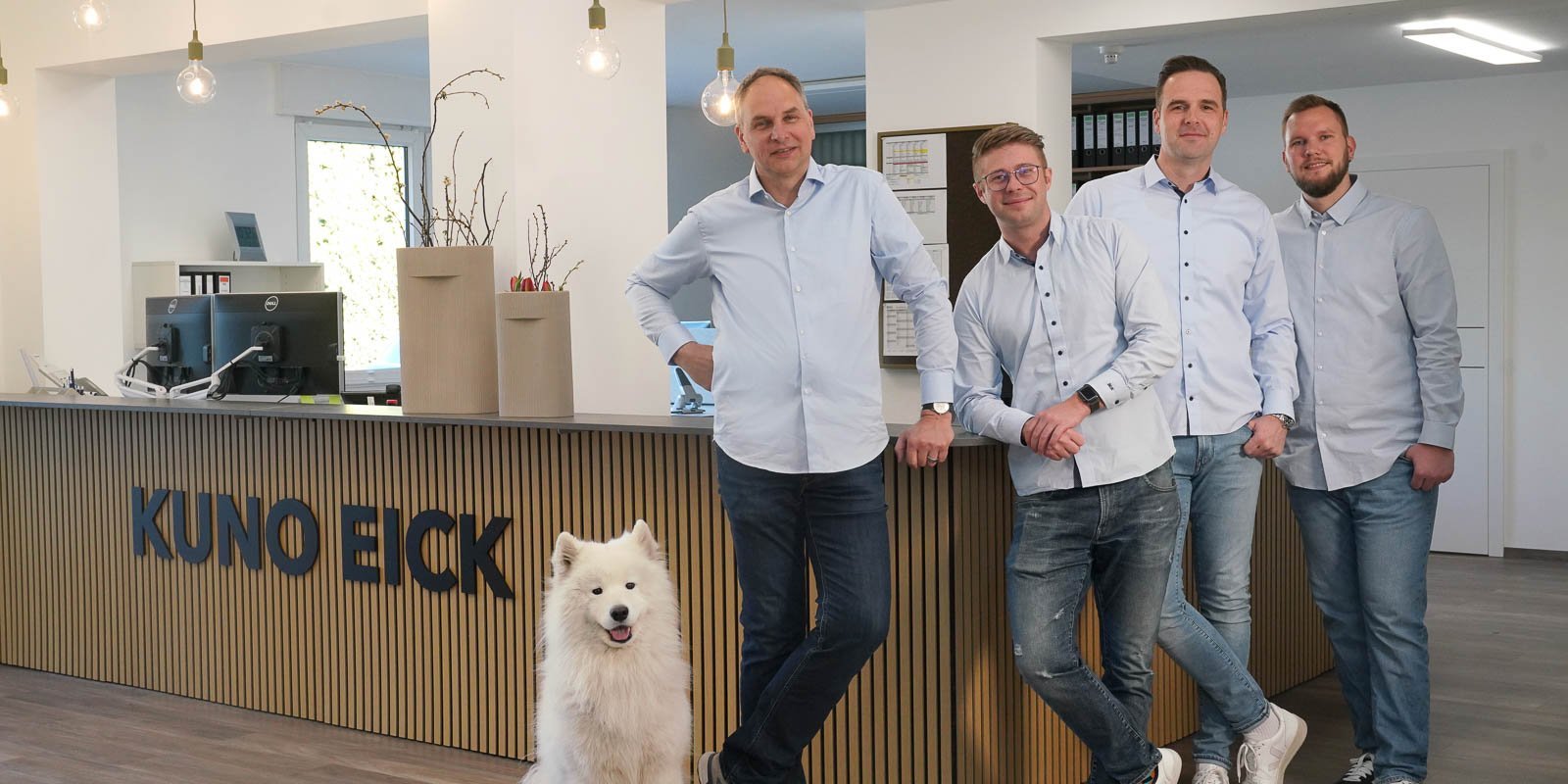In einem modernen Büro lächeln vier Männer, die in hellblauen Hemden gekleidet sind, während ein Hund vor ihnen steht.