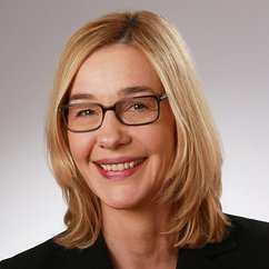 Eine lächelnde Frau mit blonden, schulterlangen Haaren und Brille, die in einem professionellen Umfeld posiert.