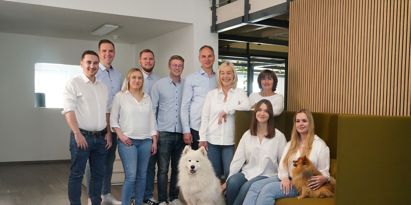 In einem hellen, modernen Büro lächeln zehn Personen, während ein flauschiger Samojede und ein kleiner Pomeranian im Vordergrund sitzen.