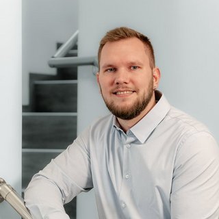 Professionelles Porträt eines lächelnden Mannes in einem modernen Raum Der Mann trägt ein hellblaues Hemd und hat einen selbstbewussten Ausdruck, während er in einem hellen Raum posiert.