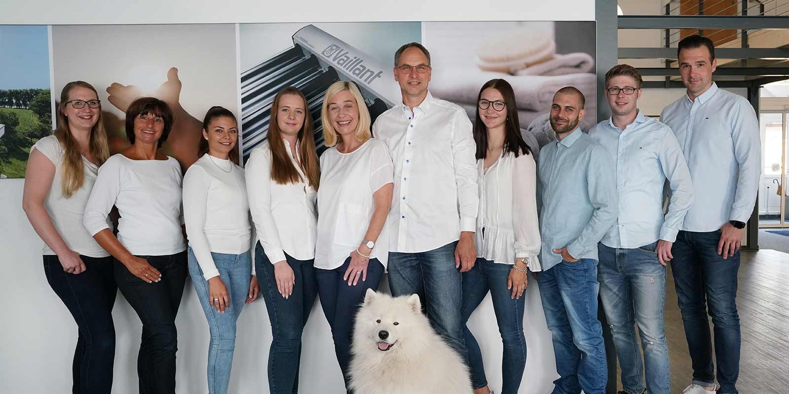 Ein Teamfoto mit lächelnden Mitarbeitern und einem Hund Die Gruppe besteht aus Männern und Frauen, die in lässiger Kleidung vor einer Wand mit einem Hund und Bildern stehen.