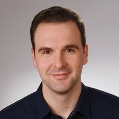 Ein junger Mann mit einem freundlichen Lächeln und kurzen, dunklen Haaren, trägt ein dunkelblaues Hemd.