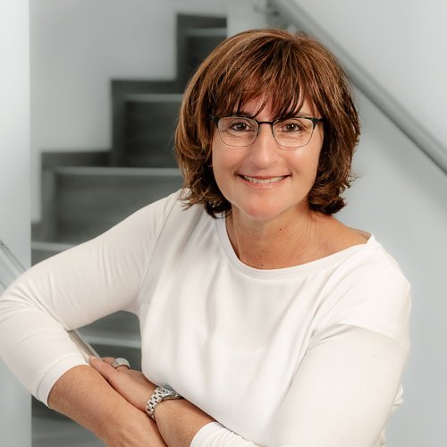 Eine lächelnde Frau mit Brille lehnt entspannt an einer Treppe und strahlt Freundlichkeit und Selbstbewusstsein aus.