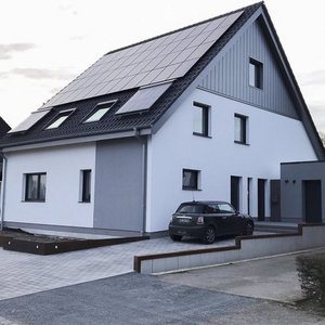 Helles Wohnhaus mit innovativer Architektur und Energieeffizienz Vor dem Haus steht ein schwarzes Auto auf einem gepflasterten Parkplatz, umgeben von einem klaren Himmel und sanften Wolken.