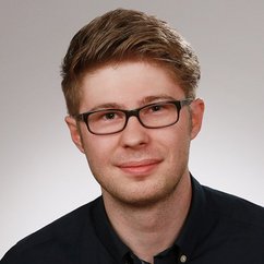 In einem neutralen Hintergrund posiert ein junger Mann mit Brille, der eine entspannte und selbstbewusste Ausstrahlung hat.