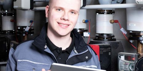 Ein junger Anlagenmechaniker vor einer Heizungsanlage, der ein Tablet hält 