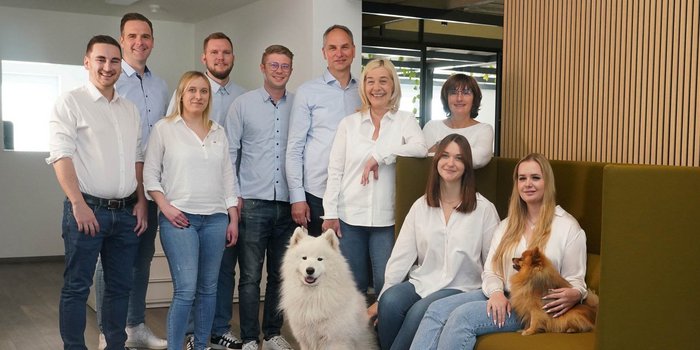 Zehn Menschen stehen und sitzen in einem hellen Raum, einige halten Hunde, während sie freundlich in die Kamera schauen.